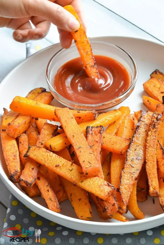 butternut squash fries in a white plate, one fries being dipped in ketchup