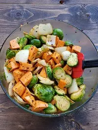 tossing sweet potatoes, brussel sprouts and onion in a clear glass bowl with a red spatula