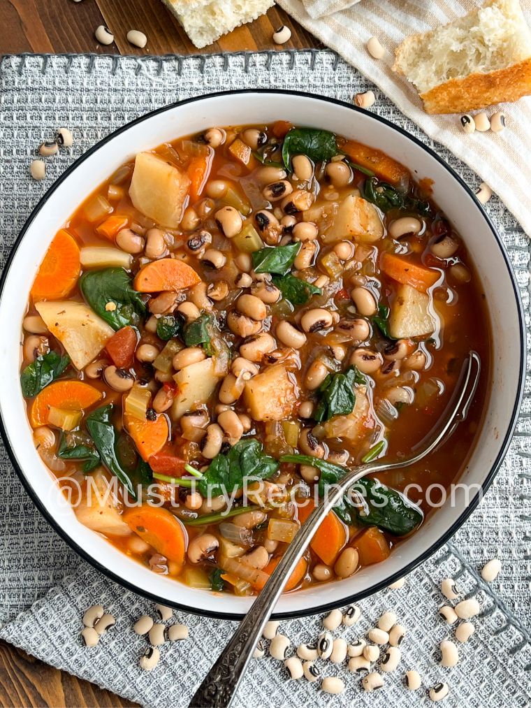 Black eyed pea soup recipe in a black rimmed white bowl with a silver spoon. The bowl in kept on a grey napkin scattered with dry black eyed peas and breads.